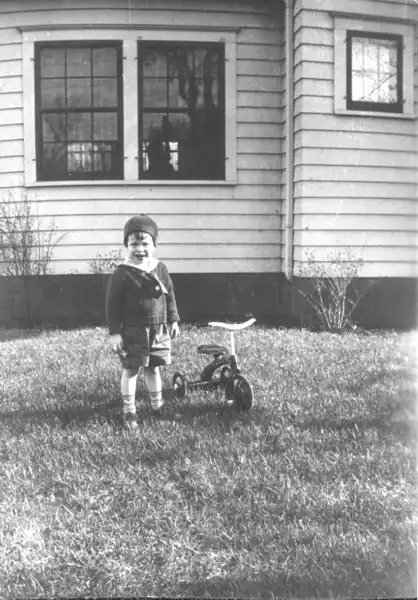 A little boy stands by a tricycle in the front yard.