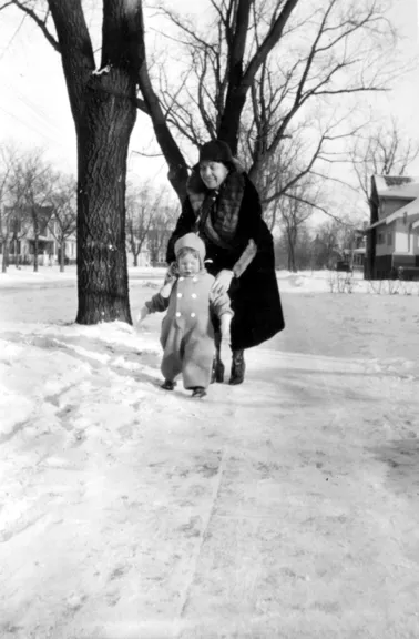 A photograph of a small boy helped by his mom to walk on a shoveled sidewalk.