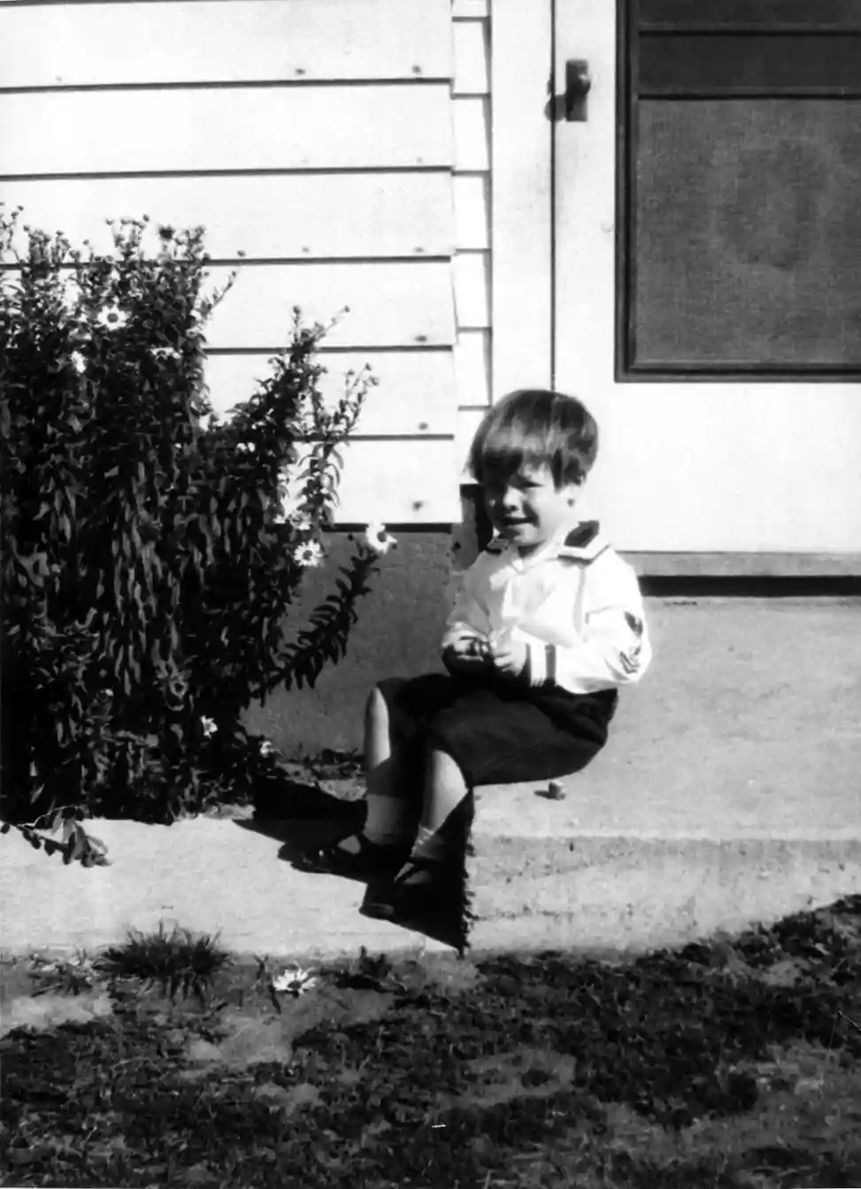 A little boy sits on a concrete step at the back door of the house.