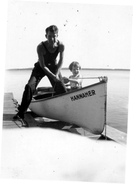 A little boy sits in a boat at the dock while a man stands.