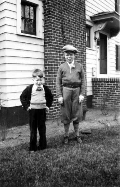A photograph of two boys standing in the front yard, both look at the camera.