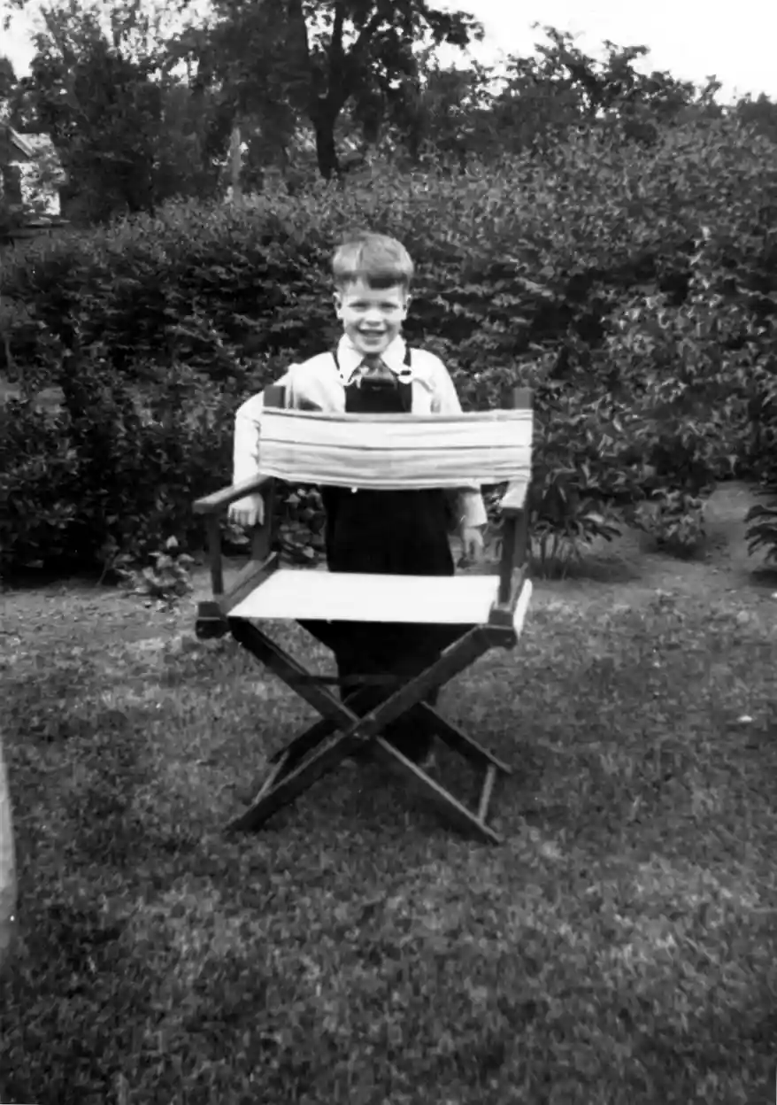 A photograph of a young boy in overalls standing in a yard behind a director's type folding chair with tall bushes behind.