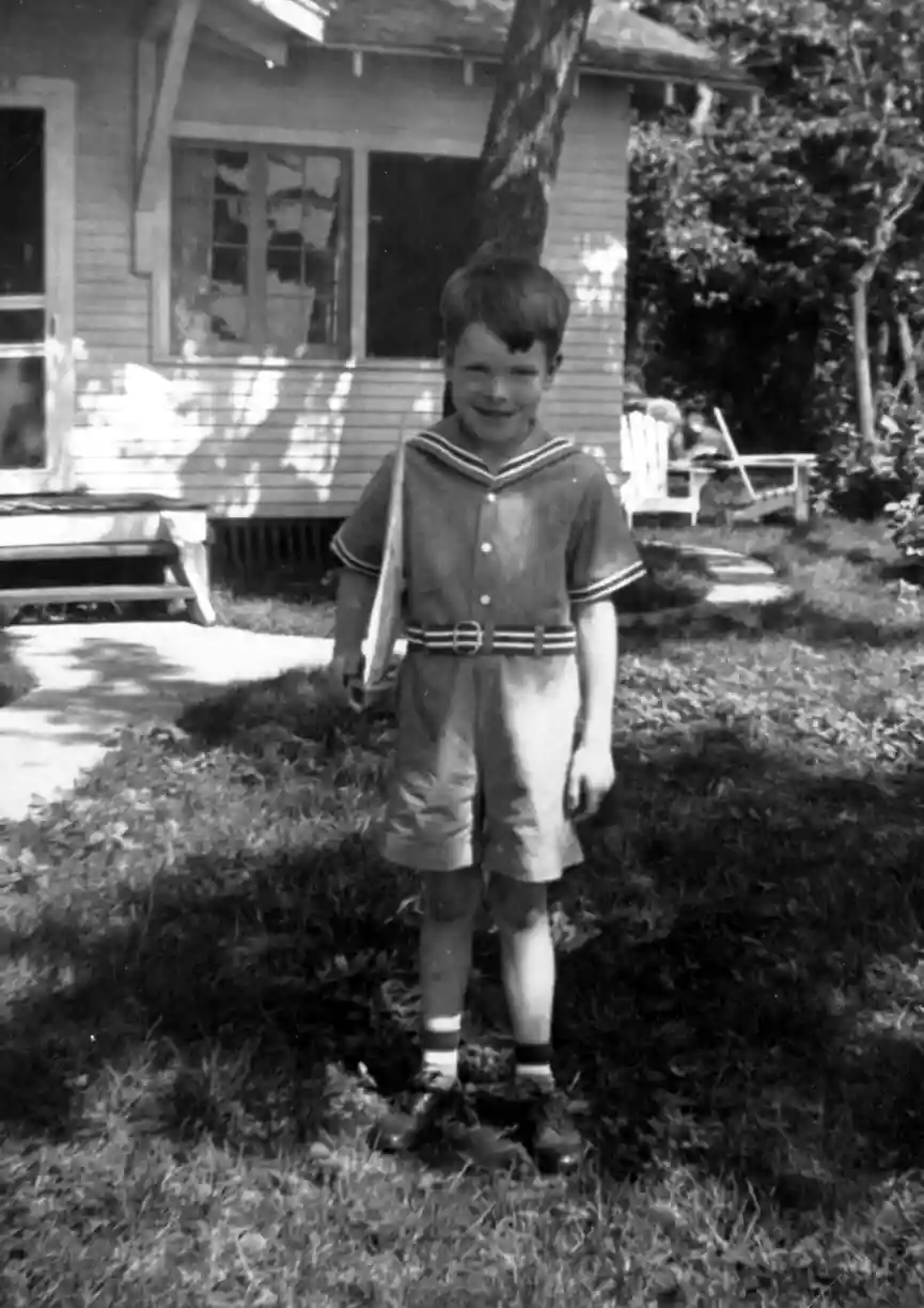 A photograph of a six year old boy standing in the grass by a lake cottage.