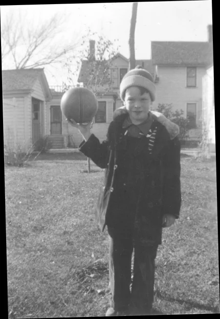 A photograph of a boy in fall clothing, holding up a ball in the backyard.