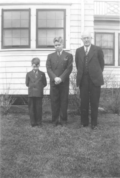 A photograph of two boys and their father, all wear suits, standing in the backyard.