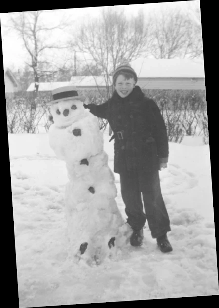 A photograph of a snowman and an eight year old boy, both are leaning.