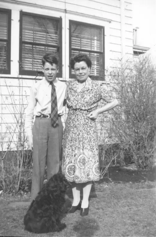 A photograph of a boy and his mother, he wears a tie. The dog has joined them in the backyard.
