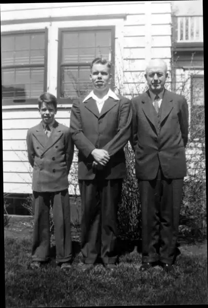 A photograph of two boys and their father standing in the backyard, all wear suits.