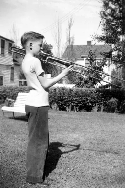 A photograph of a young man in a tee and long pants in a backyard playing a trombone.