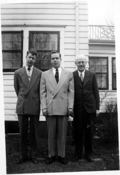 A photograph of two sons and their father, all wear suits, they are standing in the backyard.