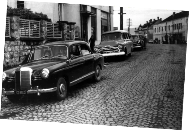 A photograph of a cobblestone street with a dark car, a truck, and another car parked.