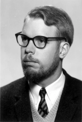 A photograph of a young bearded man wearing a suit, V-neck sweater, tie, and eyeglasses looks aside for his portrait.