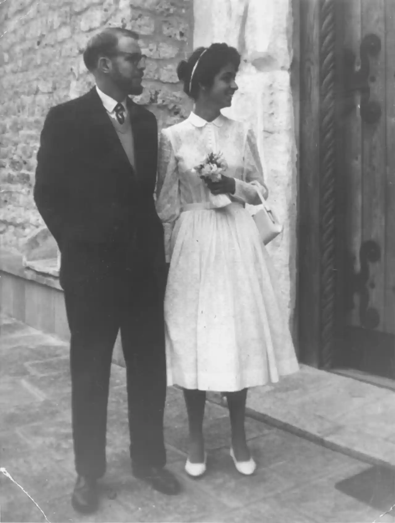 A photograph of a groom and bride standing by a heavy wooden door.