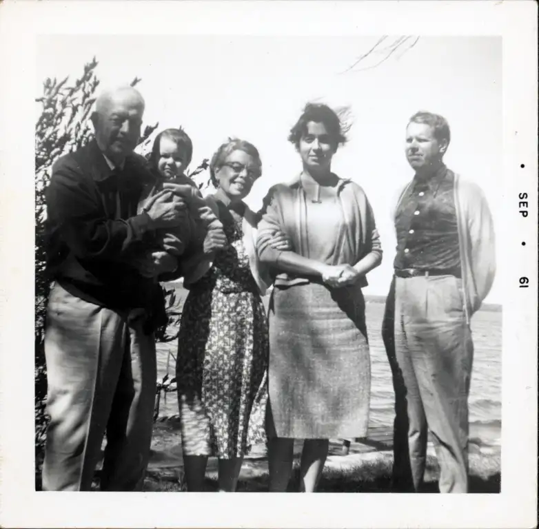 A photograph of a grandfather holding a grandson, a grandmother, a dark-haired woman, and a red-haired man at a lake shore.