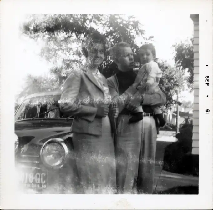 A photograph of a dark-haired woman, a red-haired man, and their son as they stand by the Mercedes-Benz 180 in the driveway in Moorhead.