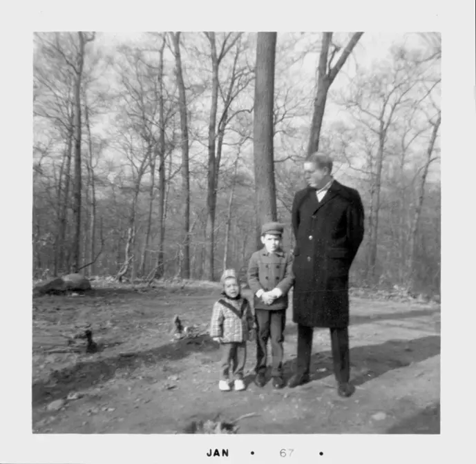 A photograph of a tall man and his two sons all in winter outerwear, in a woods.