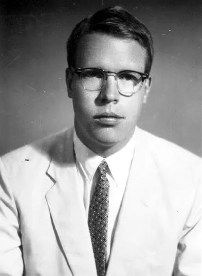 A photograph of a white man wearing a suit, tie, and eyeglasses.